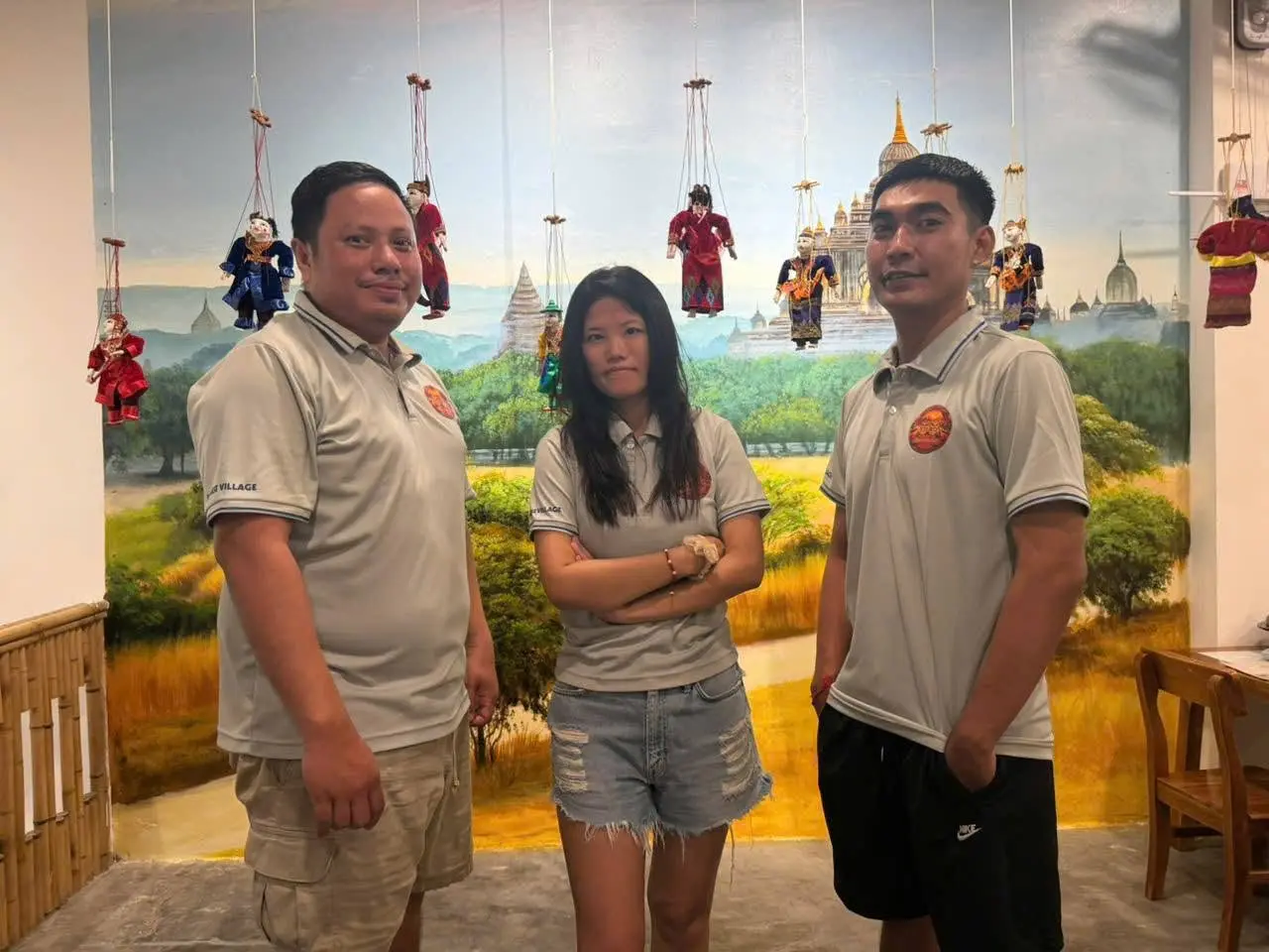 Three Burmese Village leaders in cream polo shirts standing in front of the hand-painted Bagan mural, yoke thé marionettes hanging above them